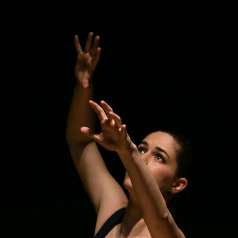A photograph of Miss Brents reaching and looking up against a dark background.