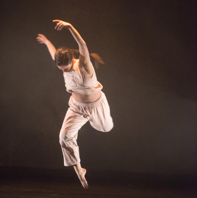 A photograph of Miss Brents leaping against a dark background. Her head is down and her arms are raised overhead. She is wearing pale beige pants and a matching shirt.